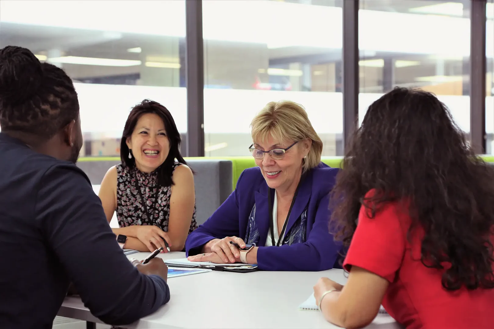 Four people seated at a conference table in a bright meeting room @ EPL, actively engaged in discussion. Two are writing notes, one uses a tablet