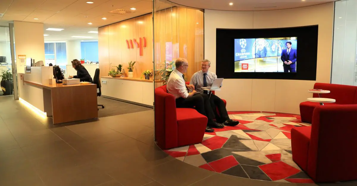 Two men in an office lobby watching a news broadcast on a large screen near a reception desk.