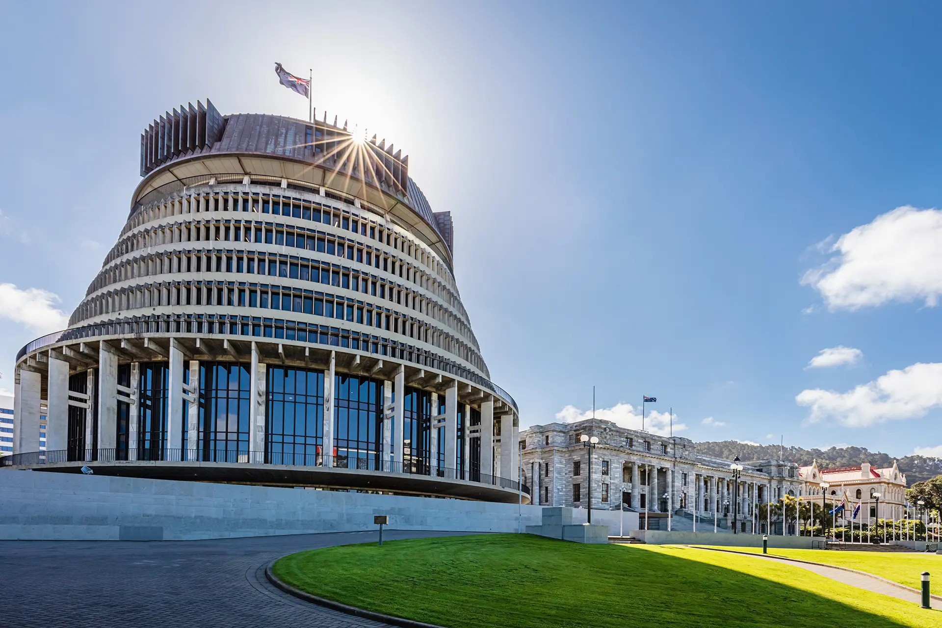 The Beehive and New Zealand Parliament Buildings in Wellington on a sunny day, with the sun shining behind the Beehive and casting rays over the modern and classical architecture.