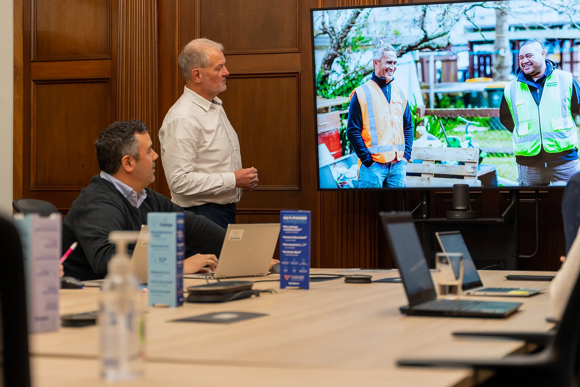 Meeting room presentation showing two construction workers on screen, with professionals seated at a table with laptops.