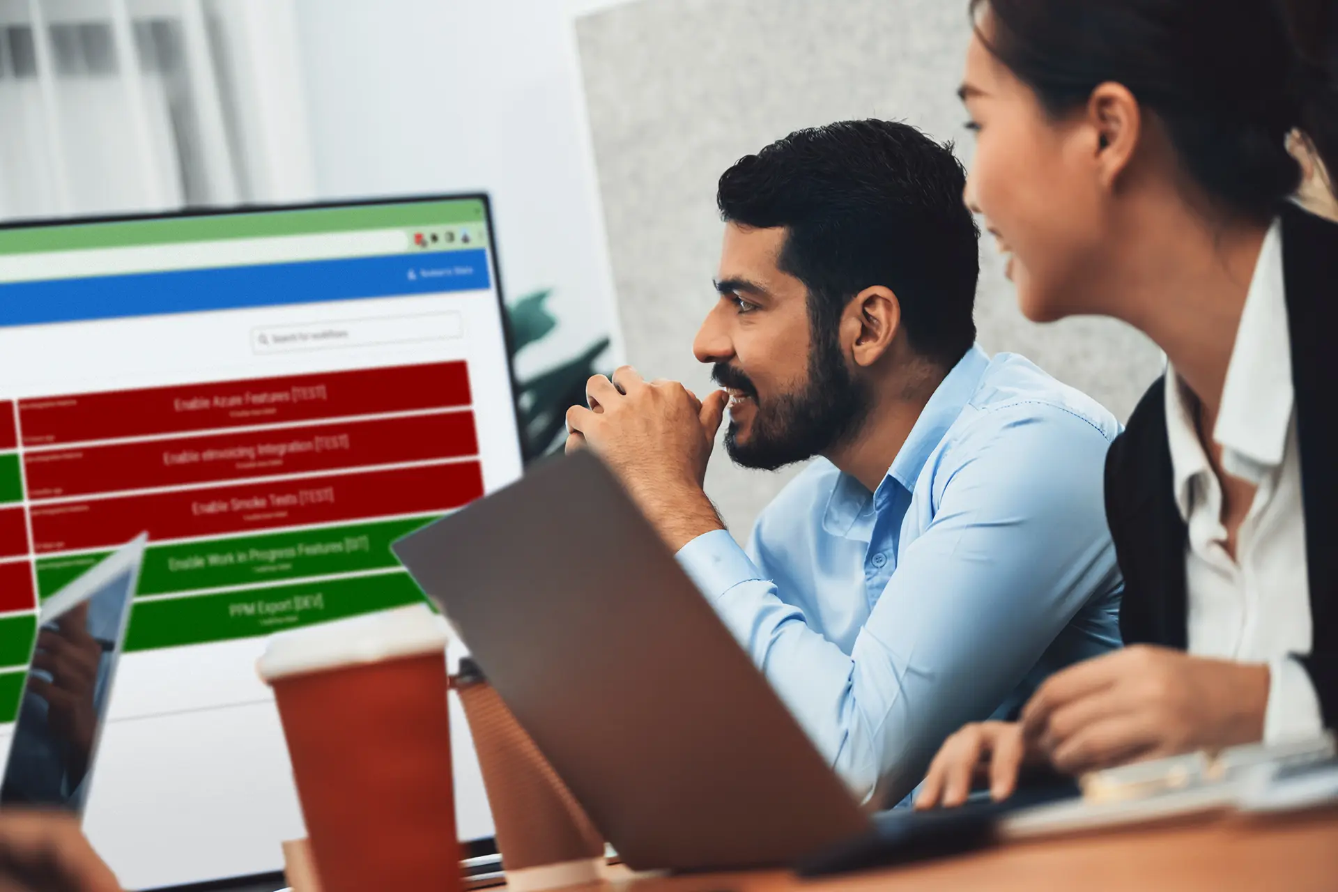 Twopeople seated at a table with laptops, viewing a large screen displaying a task list with red and green items, including Azure and Oracle features.