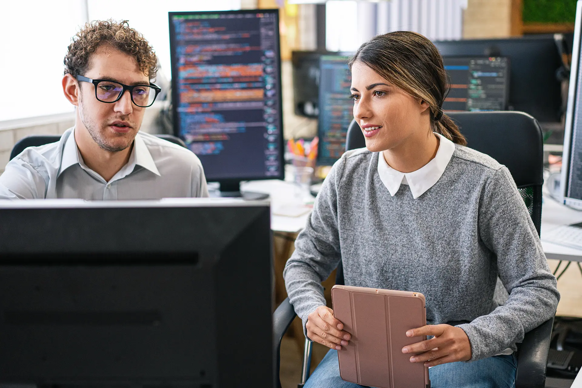 Two software testers collaborate at a desk with multiple monitors displaying code.