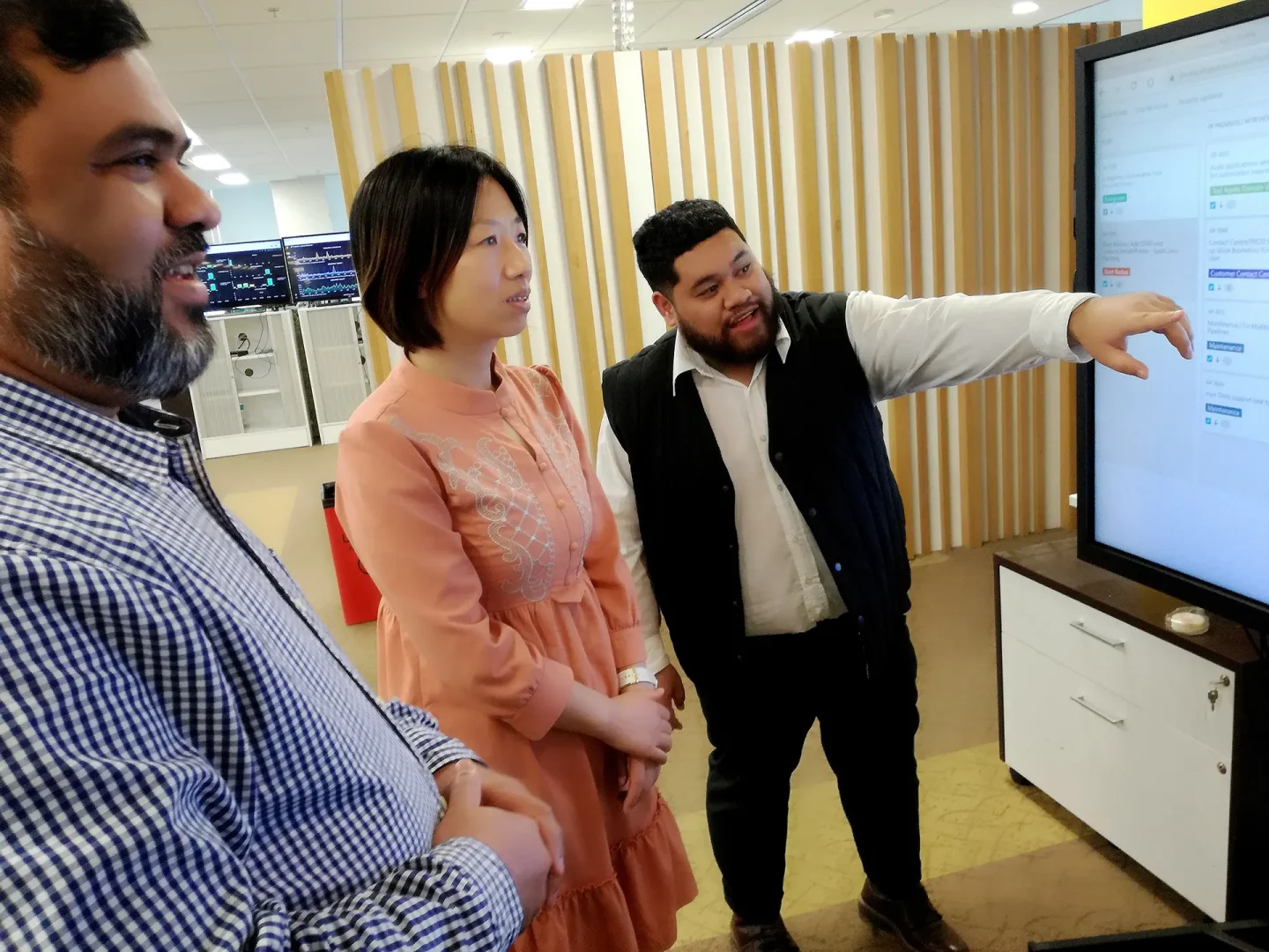 Three people in an office; one points at a large screen displaying information. Background includes a striped partition and multiple computer monitors.