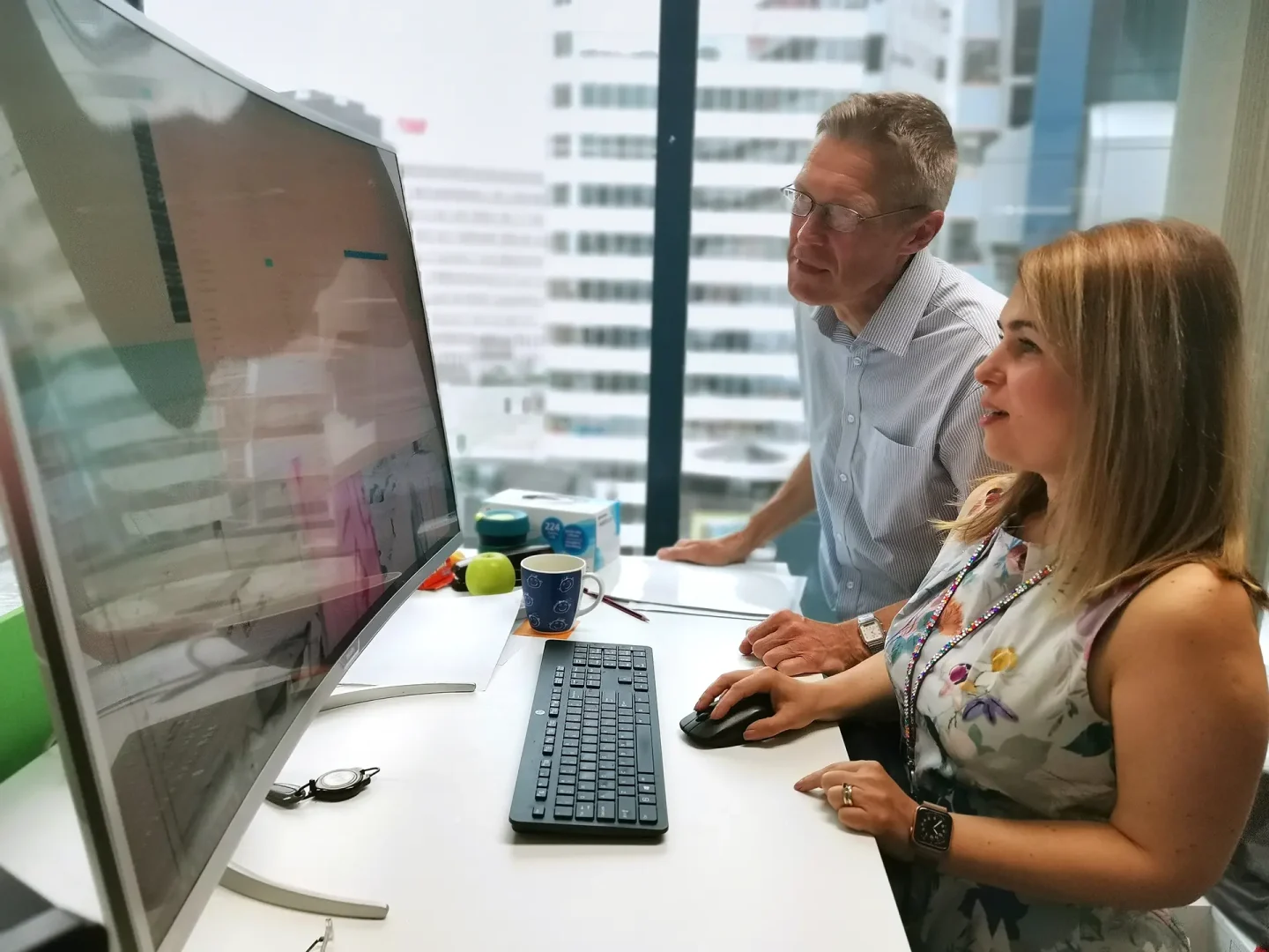 Two people seated at a desk in a modern office space , collaborating while viewing a large monitor. The scene includes a cityscape visible through large windows, with glasses, coffee cups, and documents scattered on the desk, suggesting an engaged and productive work session.