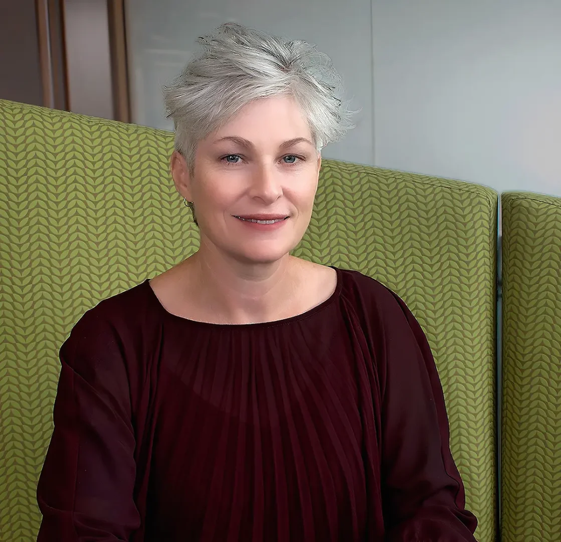 A person with short gray hair wearing a dark red top sits in front of a green patterned background. The face is blurred out.