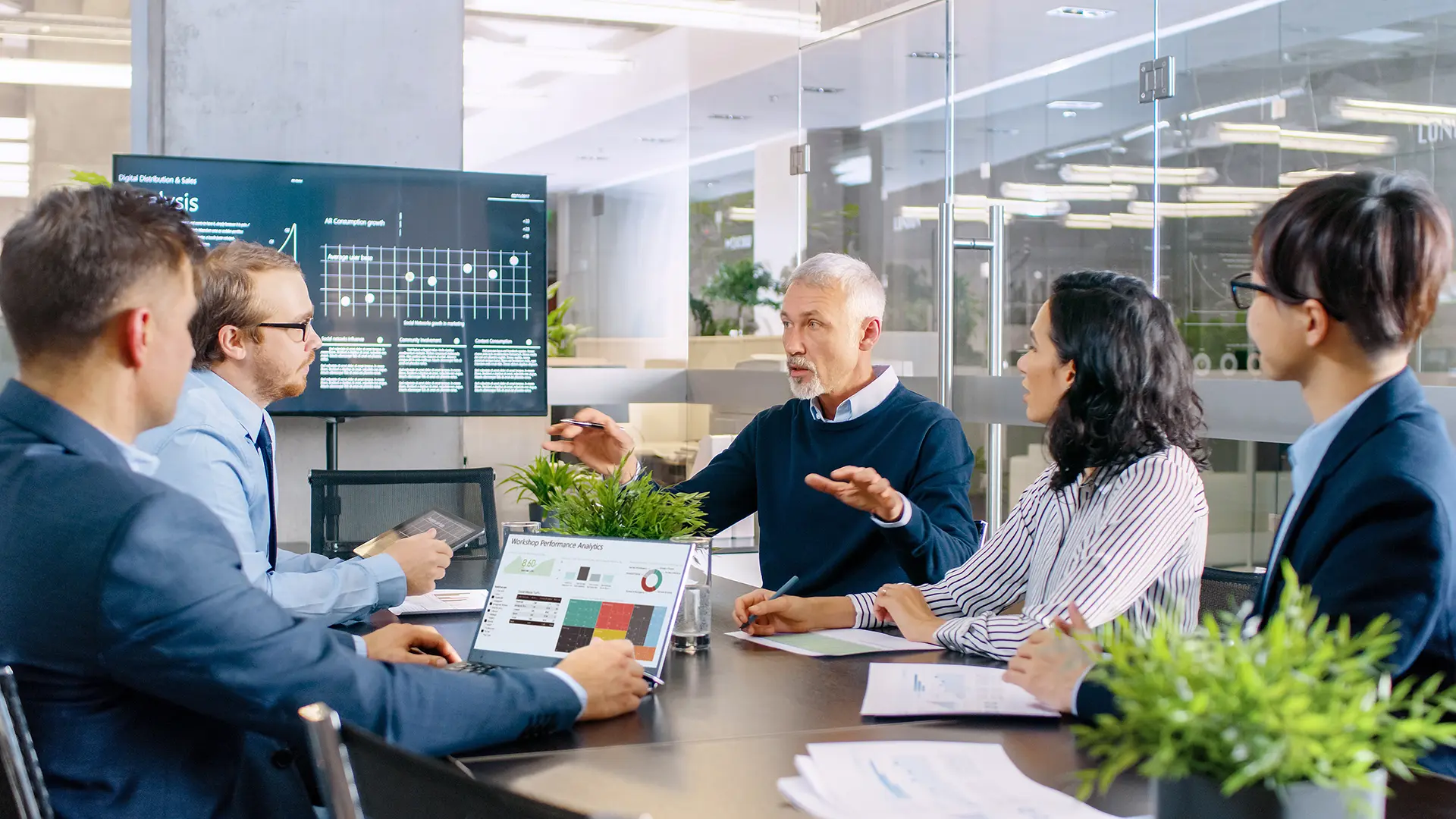 Five people in a modern office meeting room. One presents data on a large screen, another views charts on a laptop. Papers and plants are on the table.