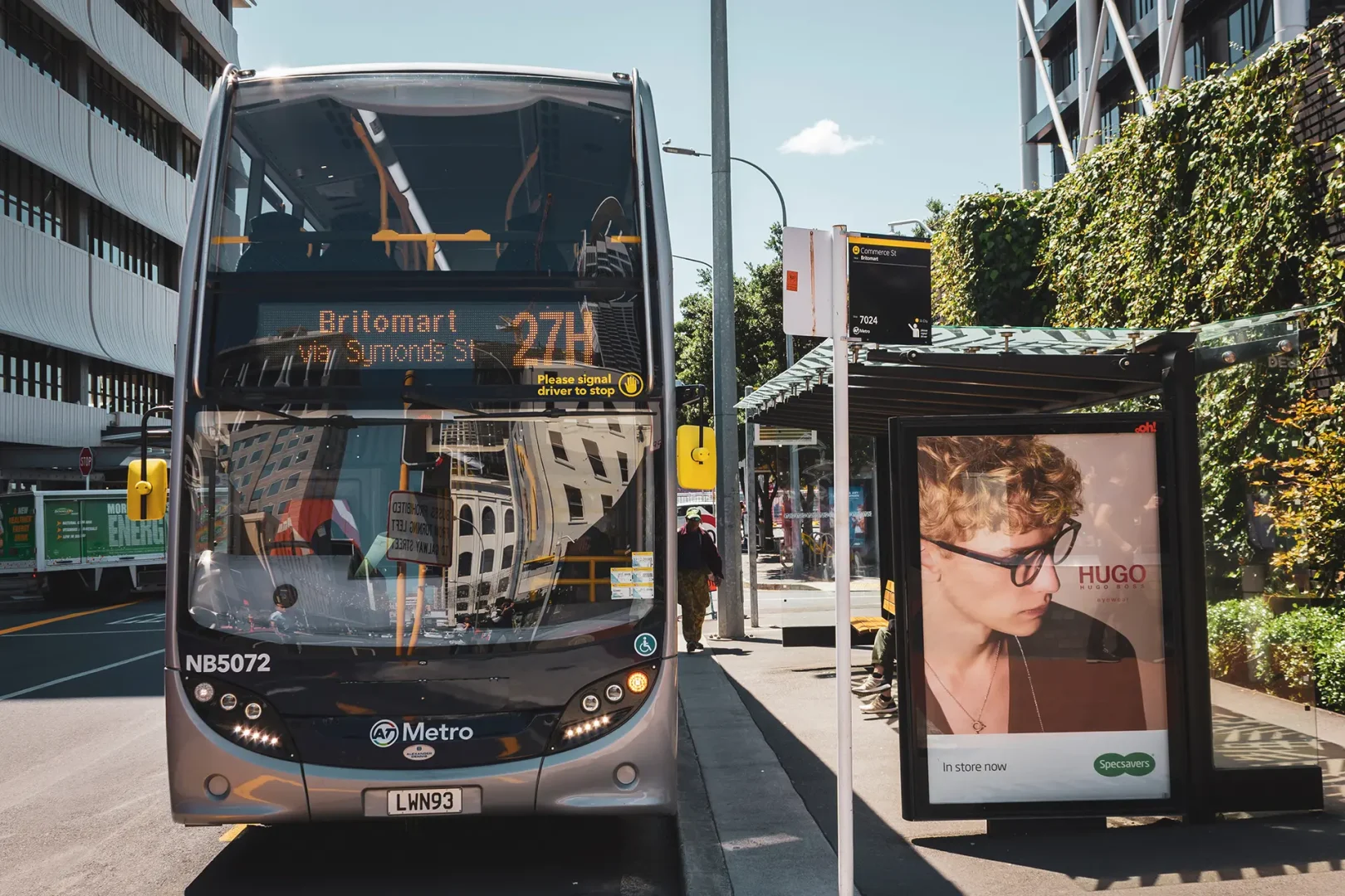 A double-decker Metro bus on route 27H to Britomart stops at a city bus stop, with pedestrians and urban buildings in the background.