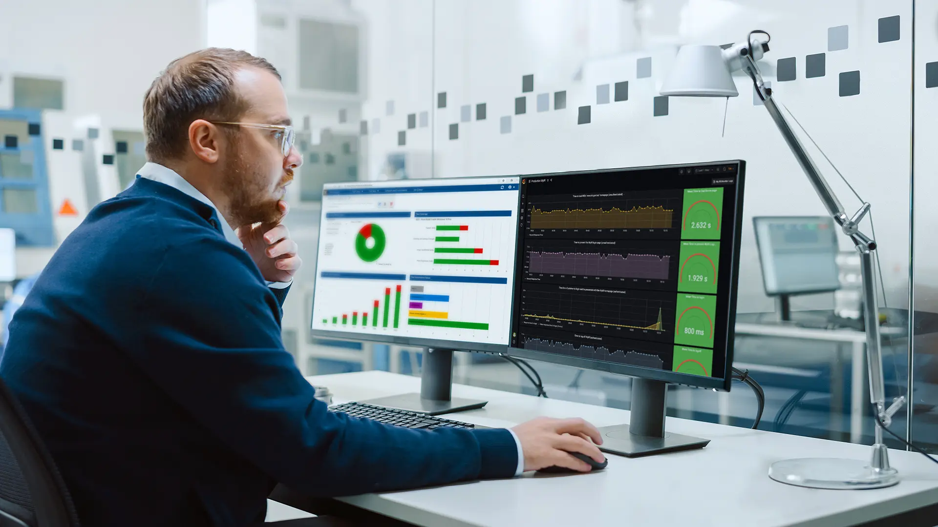 Person in a blue sweater sits at a desk in an office, viewing two computer monitors displaying charts and graphs.