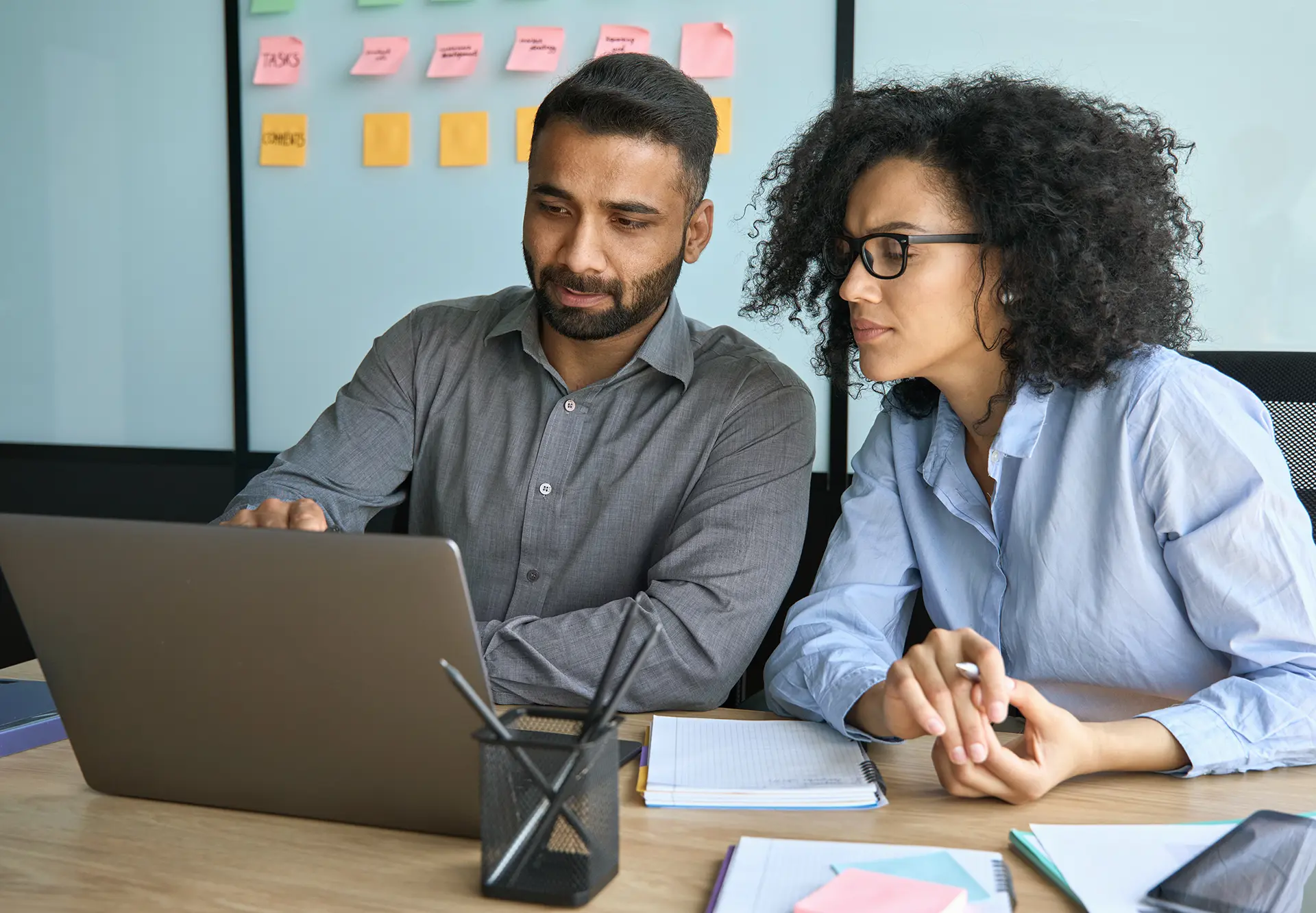 Two colleagues reviewing content on a laptop at a desk, with sticky notes on the wall behind them.