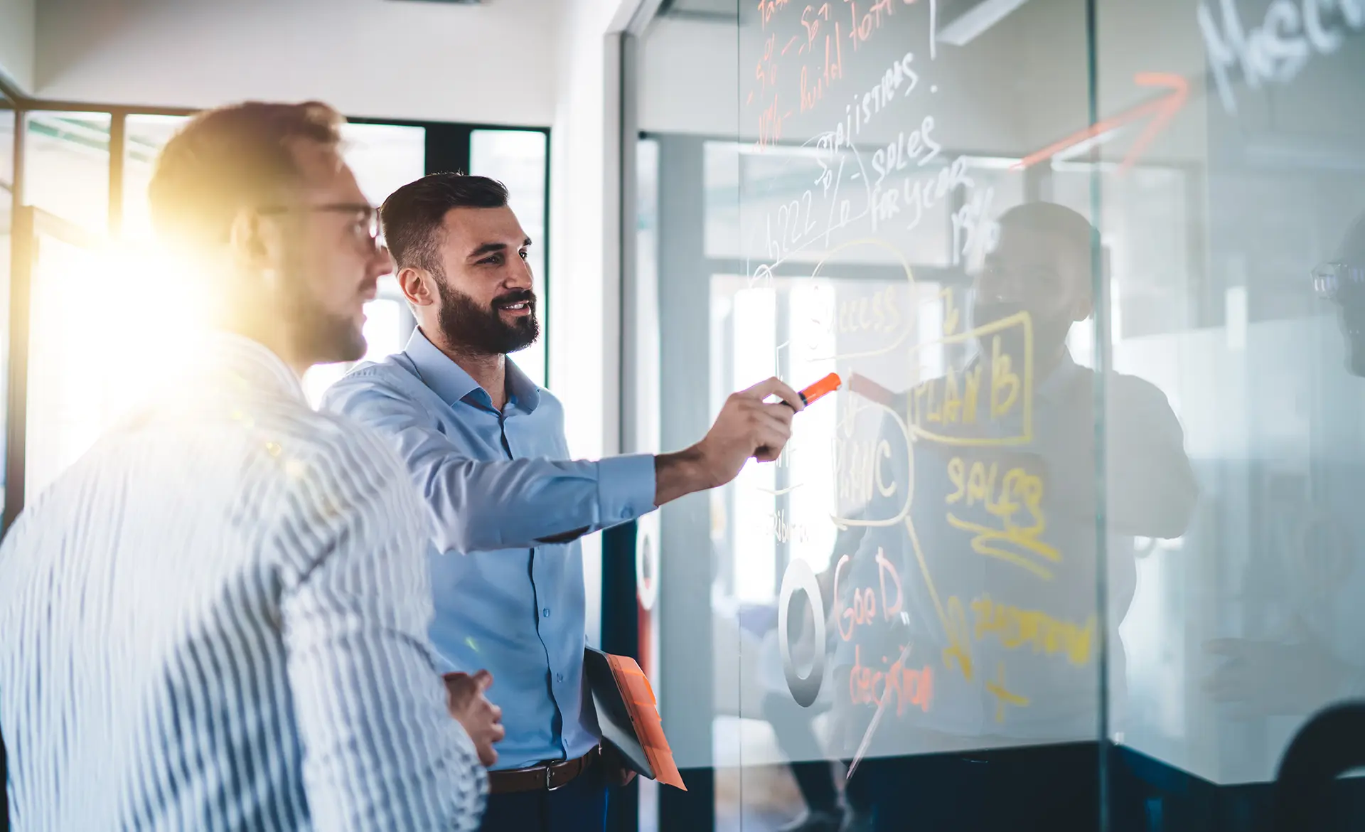 Two men collaborating and writing on a glass wall covered with colorful diagrams and notes.