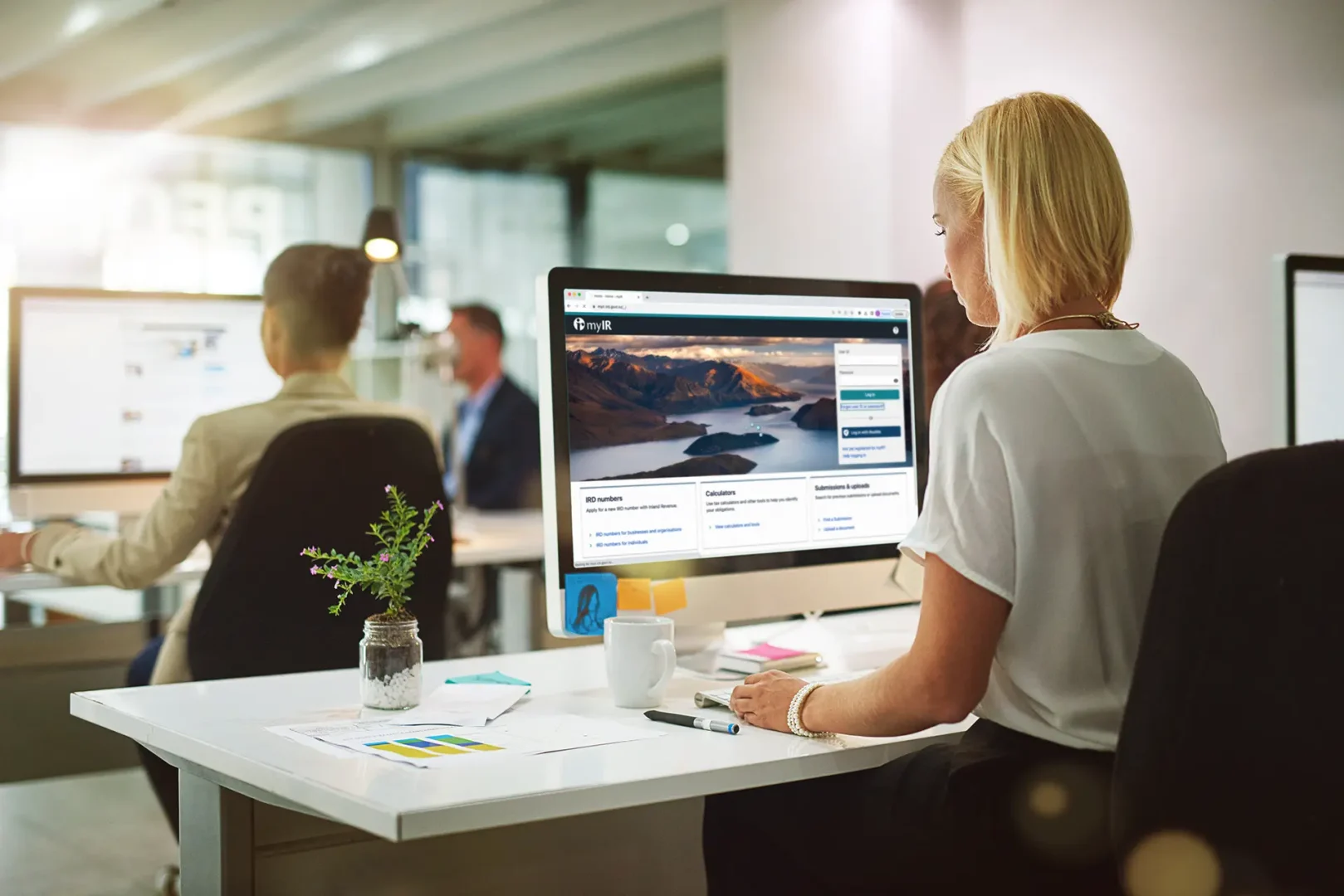 Office scene with several people at desks. A woman is the focus, working on a computer displaying a website. Desks have office supplies and decorations.