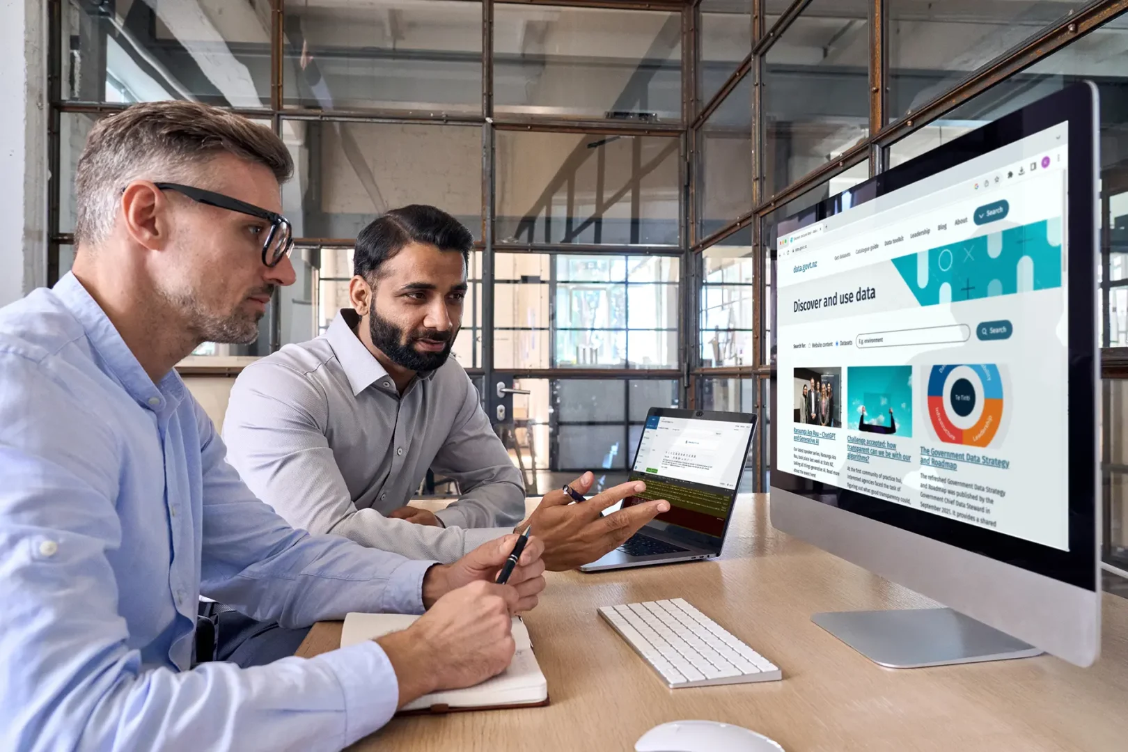 Two people in an office sit at a desk, reviewing data on a computer monitor and a laptop. One person points at the screen with a pen.