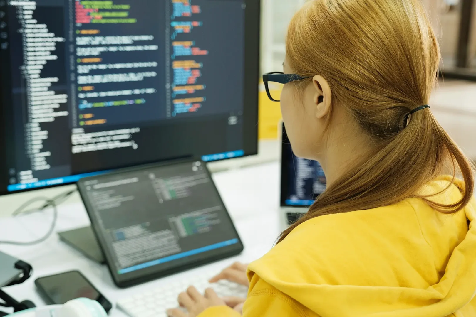 Person in a yellow hoodie working at a desk with multiple monitors displaying code. The individual's face is blurred for privacy.