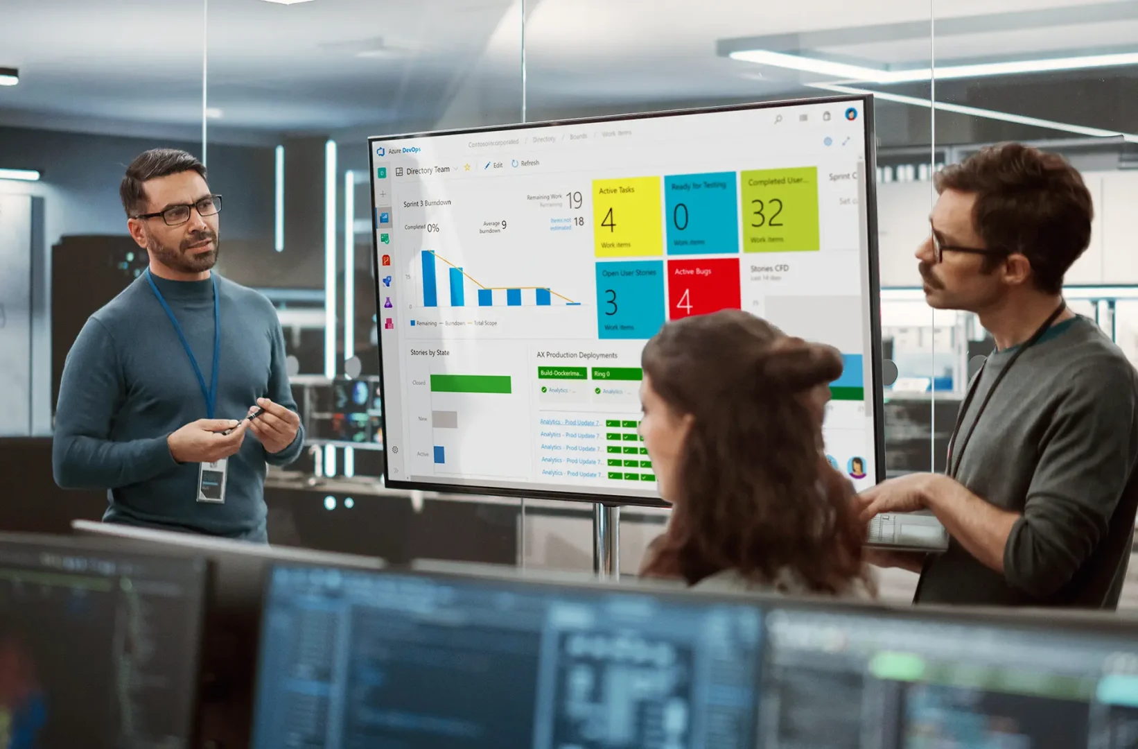 Three professionals in an office discuss project progress displayed on a large screen showing Azure DevOps dashboards with charts, task metrics, and deployment updates.