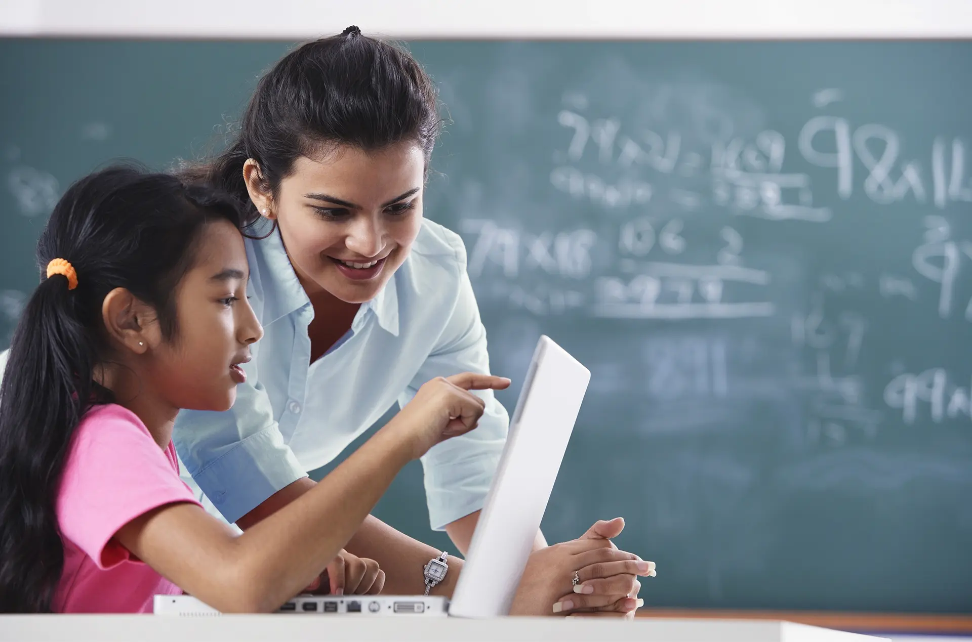 Teacher helping a student with a laptop in a classroom, with math equations on the chalkboard.