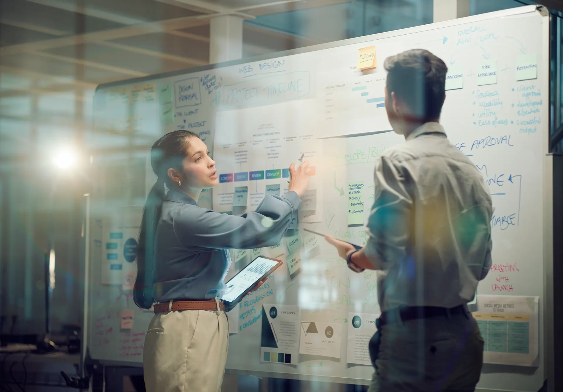 Two professionals collaborate in front of a whiteboard covered with charts, timelines, and diagrams.