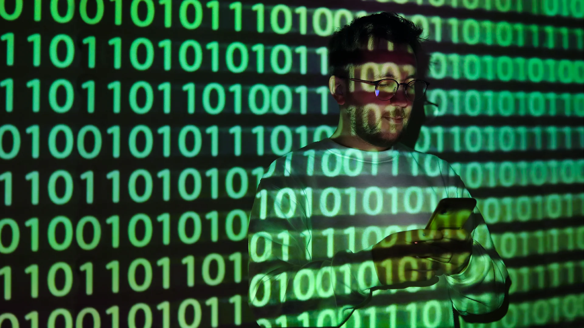 A man wearing glasses looks at a smartphone while green binary code is projected across his face, clothing, and the wall behind him, suggesting data, cybersecurity, or digital technology.